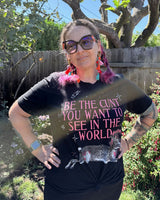 A woman wearing a black graphic t-shirt with pink text that reads 'BE THE CUNT YOU WANT TO SEE IN THE WORLD' and a rabbit illustration, posing outdoors in a garden setting.