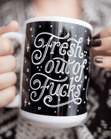 Close-up of a person holding a black and white mug with decorative text reading Fresh Out of Fucks in a cozy setting