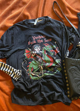 Black graphic t-shirt with punk illustration and text, paired with striped arm warmers, leather bag, spiked boots, and black pants on orange background